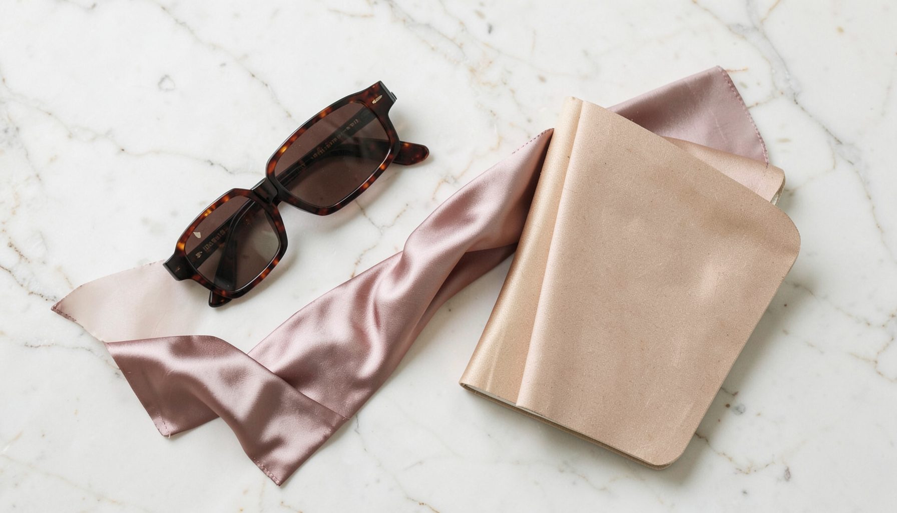 A flat lay of fashion accessories — sunglasses, a silk scarf, and a notebook — on a pale marble surface, soft natural light