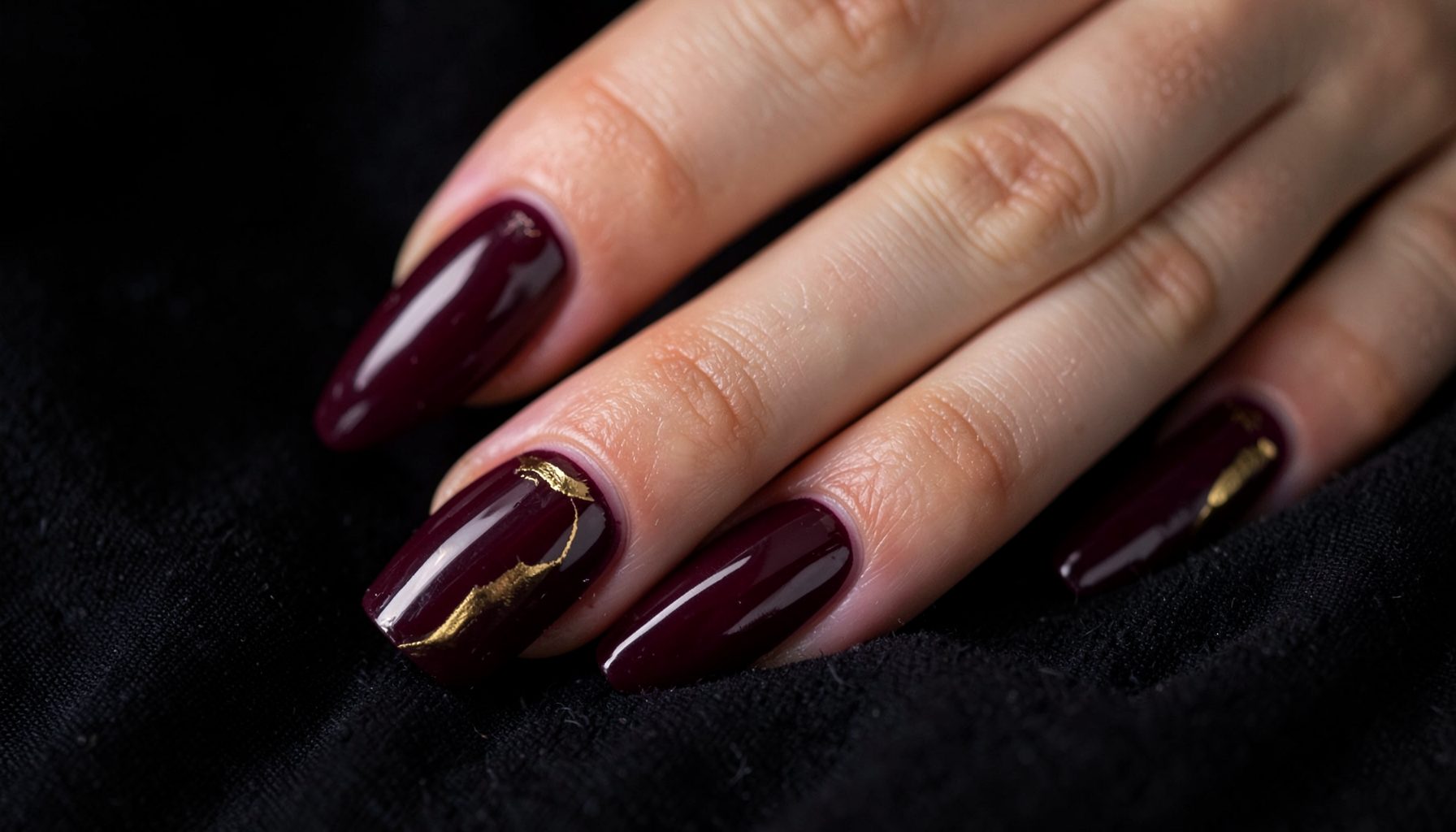 Close-up of glossy nails in deep burgundy and gold tones against a dark fabric background, moody and luxurious